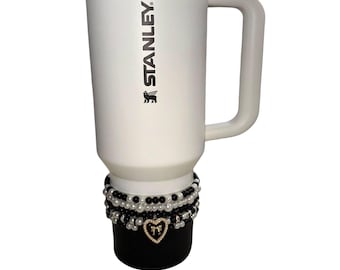 May include: White Stanley travel mug with a black lid and handle. The mug has the Stanley logo printed in black. The base of the mug is decorated with multiple strands of black and white beads and a gold heart charm.
