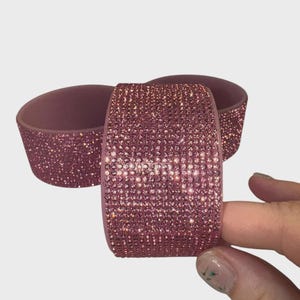 May include: Three mauve-colored, bedazzled bracelets. One bracelet is held by a hand. The bracelets are covered in small, sparkling pink rhinestones. The background is a plain, light gray.