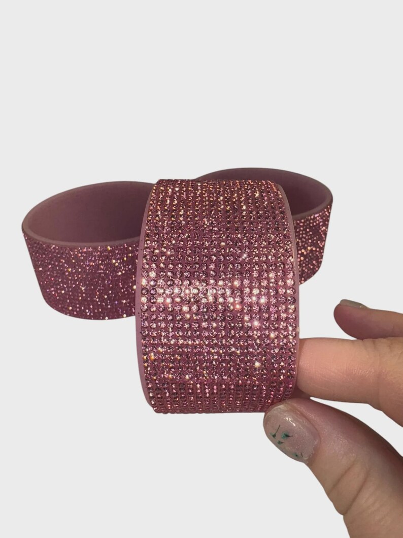 May include: Three mauve-colored, bedazzled bracelets. One bracelet is held by a hand. The bracelets are covered in small, sparkling pink rhinestones. The background is a plain, light gray.