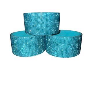 Puede incluir: Tres fundas para bebidas de color turquesa. Cada funda cil&iacute;ndrica est&aacute; cubierta en el exterior con brillantes pedrer&iacute;a iridiscente. El interior de cada funda es de color turquesa liso. Las fundas est&aacute;n apiladas en forma triangular.