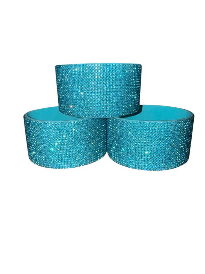 Puede incluir: Tres fundas para bebidas de color turquesa. Cada funda cil&iacute;ndrica est&aacute; cubierta en el exterior con brillantes pedrer&iacute;a iridiscente. El interior de cada funda es de color turquesa liso. Las fundas est&aacute;n apiladas en forma triangular.
