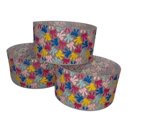 Puede incluir: Tres recipientes cilíndricos decorativos. Cada recipiente está adornado con un patrón de lazos coloridos en rosa, amarillo, azul y rosa claro. Los recipientes están dispuestos en una configuración apilada, mostrando su diseño.