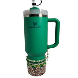 May include: A green Stanley travel mug with a gold chain and a green and white beaded bracelet. The mug has a clear lid with a straw and a green clover charm.