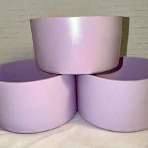 May include: Three light purple, cylindrical containers are stacked together. The containers have a smooth, matte finish and a rounded bottom. The top container has a subtle iridescent sheen. The containers are likely designed to hold items.