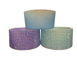 May include: Three decorative drink sleeves in pastel colors. The top sleeve is light blue with glitter, while the bottom sleeves are purple and teal, each covered in sparkling rhinestones. These sleeves are designed to insulate beverages.