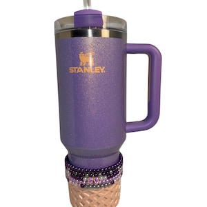Op de afbeelding: Paarse reistumbler van roestvrij staal met een handvat en een rietjesdeksel. De tumbler heeft een glitterafwerking en het Stanley-logo is aan de zijkant gedrukt. De tumbler is versierd met een roze en paarse kralenwikkeling.