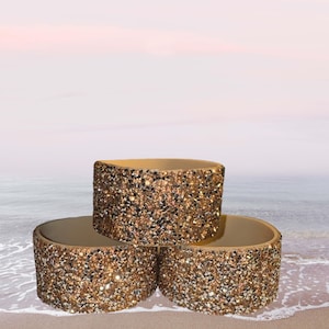 Puede incluir: Tres recipientes decorativos cilíndricos con una superficie brillante y texturizada en tonos dorados y bronce. Los recipientes están apilados, con los dos inferiores descansando sobre una superficie arenosa, y el superior ligeramente elevado. El fondo presenta una escena de playa.