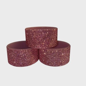 May include: Three stacked, cylindrical napkin rings. Each ring is covered in small, sparkling, pink rhinestones. The napkin rings are arranged in a triangular formation against a plain white background.