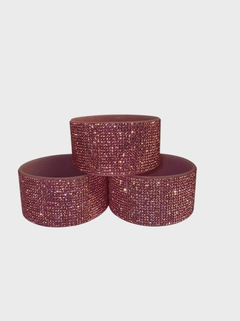 May include: Three stacked, cylindrical napkin rings. Each ring is covered in small, sparkling, pink rhinestones. The napkin rings are arranged in a triangular formation against a plain white background.