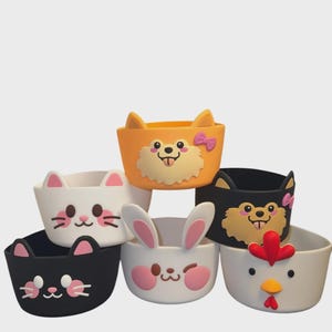 May include: Six animal-shaped containers in a variety of colours. The containers are designed to resemble a cat, dog, bunny, and chicken. The colours include orange, white, black, and pink. The containers are made from a soft, flexible material.