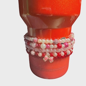 Op de afbeelding: Een rode glitterbeker met een stapel kralenarmbanden in tinten roze, rood en wit. De armbanden hebben parels en decoratieve kralen, met een roze strikbedel. Drinkware accessoire.