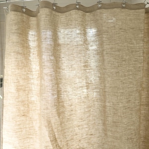 Puede incluir: Una cortina de ducha de lino beige cuelga de una barra de metal. La cortina tiene un aspecto texturizado y es ligeramente transl&uacute;cida, permitiendo el paso de la luz. El borde superior est&aacute; reforzado con una banda m&aacute;s oscura y ojales met&aacute;licos.