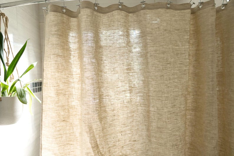 Puede incluir: Una cortina de ducha de lino beige cuelga de una barra de metal. La cortina tiene un aspecto texturizado y es ligeramente transl&uacute;cida, permitiendo el paso de la luz. El borde superior est&aacute; reforzado con una banda m&aacute;s oscura y ojales met&aacute;licos.