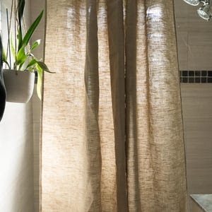 Puede incluir: Una cortina de ducha de lino beige cuelga de una barra de metal curvada. La cortina tiene un aspecto texturizado y est&aacute; parcialmente corrida, revelando un vistazo a la zona de la ducha. Se ve una planta colgante en una maceta blanca.