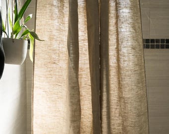 May include: A neutral-toned linen shower curtain hangs from a curved metal rod. The curtain has a textured appearance and is partially drawn, revealing a glimpse of the shower area. A hanging plant in a white pot is visible.