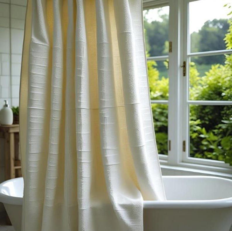 Puede incluir: Cortina de ducha color crema con un patr&oacute;n de rayas verticales texturizadas. La cortina cuelga frente a una ba&ntilde;era blanca, con una ventana y vegetaci&oacute;n visible en el fondo. La cortina est&aacute; hecha de una tela suave y fluida.