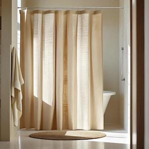 Puede incluir: Una cortina de ducha beige cuelga de una barra plateada en un ba&ntilde;o. La cortina tiene rayas verticales de diferentes tonos de beige. Una toalla a juego cuelga de la pared. Una alfombra redonda y tejida est&aacute; en el suelo.