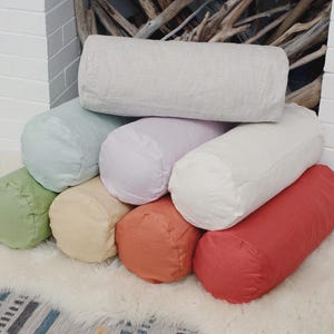 May include: A collection of colorful bolster pillows in various shades, including white, green, pink, orange, and red. The cylindrical pillows are stacked on a white, fluffy surface, creating a visually appealing display.