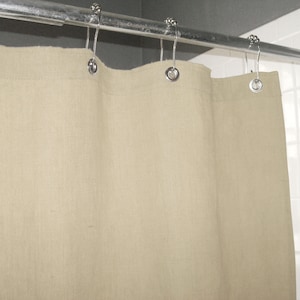 Puede incluir: Una cortina de ducha de lino beige cuelga de una barra de metal plateado con ganchos decorativos. La tela texturizada de la cortina forma pliegues suaves.
