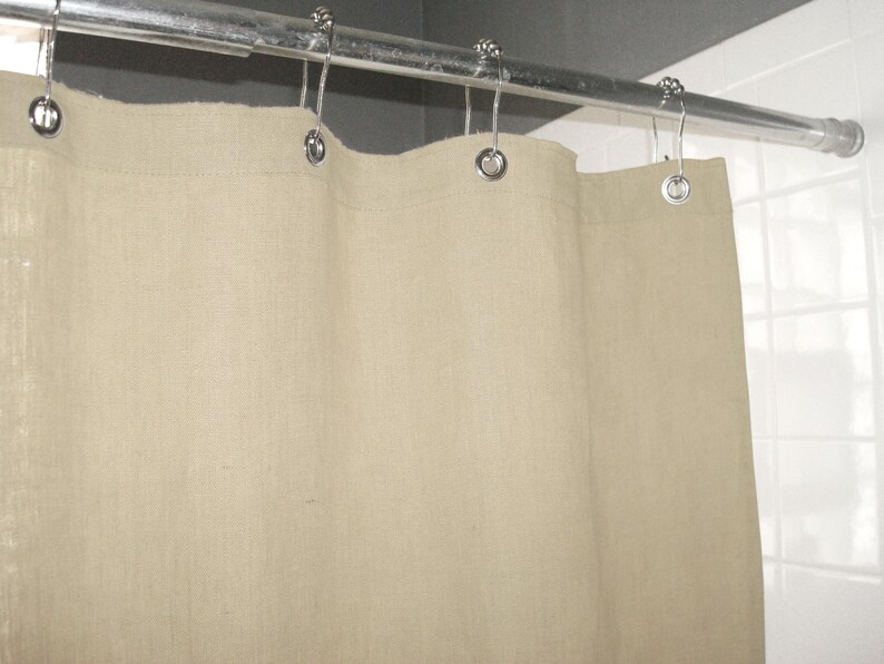 Puede incluir: Una cortina de ducha de lino beige cuelga de una barra de metal plateado con ganchos decorativos. La tela texturizada de la cortina forma pliegues suaves.