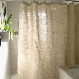 Puede incluir: Una cortina de ducha de lino beige cuelga de una barra de metal curvada en un ba&ntilde;o. La cortina tiene un aspecto texturizado y cuelga en pliegues suaves. Se ve un mueble blanco con encimera de m&aacute;rmol.