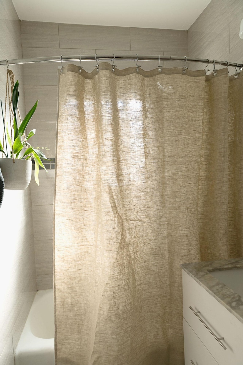 Puede incluir: Una cortina de ducha de lino beige cuelga de una barra de metal curvada en un ba&ntilde;o. La cortina tiene un aspecto texturizado y cuelga en pliegues suaves. Se ve un mueble blanco con encimera de m&aacute;rmol.