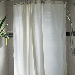 Puede incluir: Una cortina de ducha blanca cuelga de una barra curva plateada en un ba&ntilde;o. La cortina est&aacute; hecha de una tela de color claro y est&aacute; parcialmente cerrada. Las paredes del ba&ntilde;o est&aacute;n alicatadas en un color gris claro.