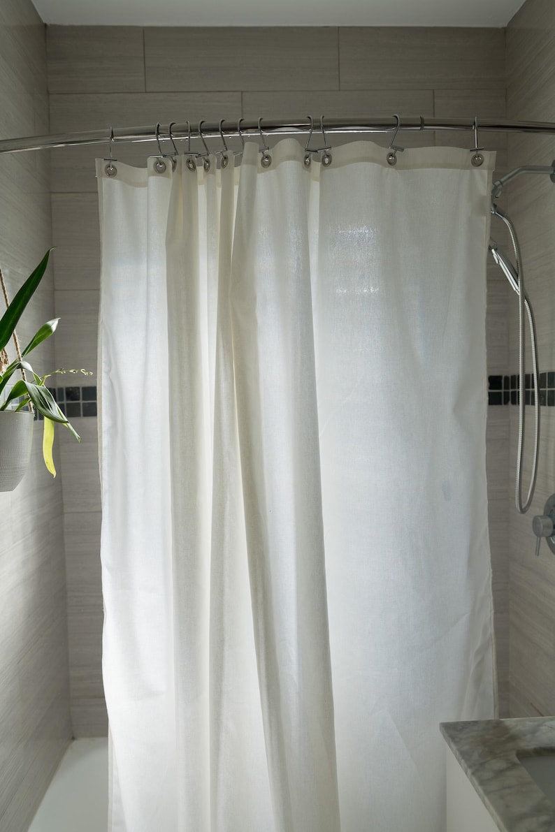 Puede incluir: Una cortina de ducha blanca cuelga de una barra curva plateada en un ba&ntilde;o. La cortina est&aacute; hecha de una tela de color claro y est&aacute; parcialmente cerrada. Las paredes del ba&ntilde;o est&aacute;n alicatadas en un color gris claro.