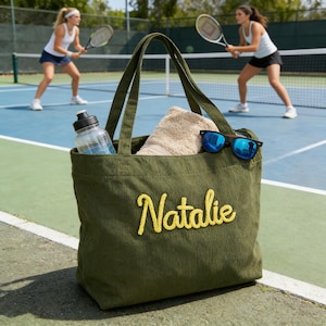 以下が含まれることがあります： オリーブグリーンのコーデュロイのトートバッグ。黄色で「Natalie」と刺繍されています。バッグには、水筒、ベージュのタオル、青いサングラスが入っています。背景には、青いコートでテニスをする2人の選手がいます。