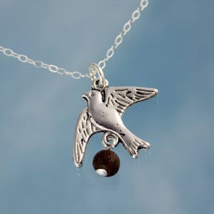 May include: A silver necklace featuring a delicate silver bird pendant and a brown wooden bead. The bird is in flight with its wings outstretched.