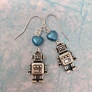May include: Silver robot earrings with light blue heart charms. The robots have a silver finish and have a simple design with a round head and a rectangular body.