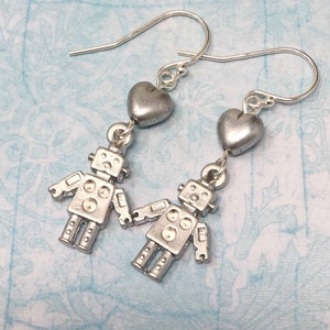 May include: A pair of silver robot earrings with heart charms. The robots are silver and have a detailed design. The hearts are also silver and have a smooth finish.