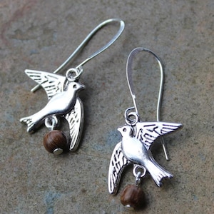 Könnte beinhalten: Silberne Taubenohrringe mit braunen Holzperlen-Akzenten. Die Tauben sind im Flug mit ausgebreiteten Flügeln.