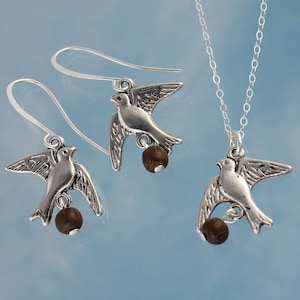 May include: A silver necklace and earrings set featuring a delicate bird charm with a brown bead. The bird is in flight with its wings spread.