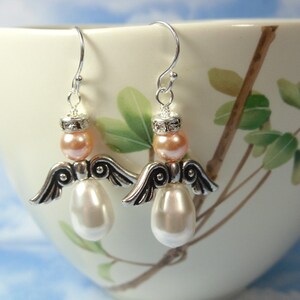 May include: A pair of angel-shaped earrings. Each earring features a silver-tone metal wing design, a peach-colored bead, and a large, pearlescent white bead. The earrings hang from silver-tone ear wires. The background is a white teacup with a floral design.