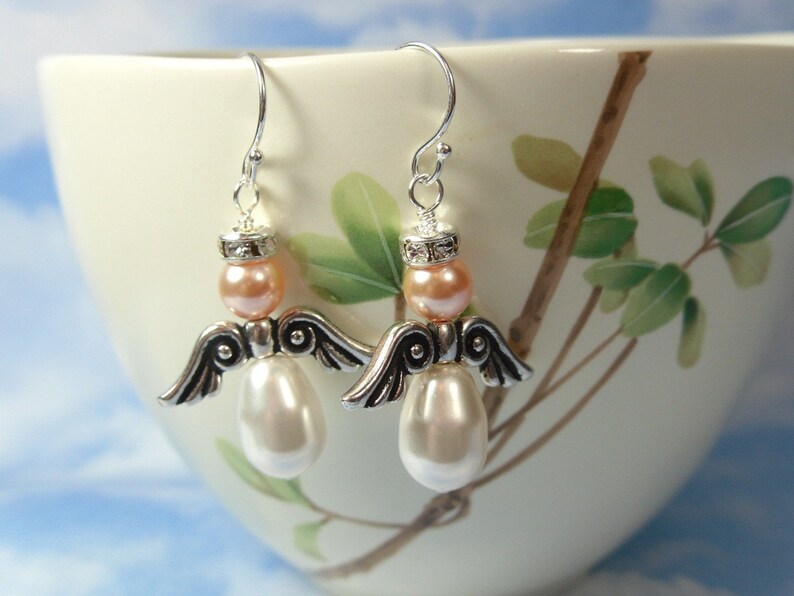 May include: A pair of angel-shaped earrings. Each earring features a silver-tone metal wing design, a peach-colored bead, and a large, pearlescent white bead. The earrings hang from silver-tone ear wires. The background is a white teacup with a floral design.