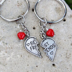May include: Two silver keychains with red heart charms. The keychains feature two halves of a heart with the words "Best Friends" on them.