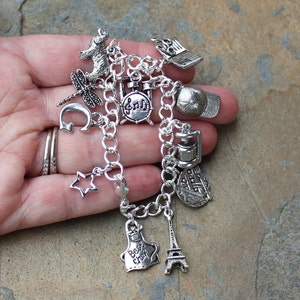May include: A silver charm bracelet featuring a range of charms, including a drum set, a star, a chef's hat, a lighthouse, an Eiffel Tower, a dragonfly, a bear, a baseball cap, and a music note.
