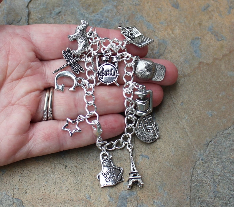 May include: A silver charm bracelet featuring a range of charms, including a drum set, a star, a chef's hat, a lighthouse, an Eiffel Tower, a dragonfly, a bear, a baseball cap, and a music note.