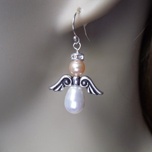 May include: A silver-toned angel earring with a peach-colored bead head, a white pear-shaped pearl body, and silver wings. The earring is suspended from a silver hook, showcasing a delicate and elegant design.