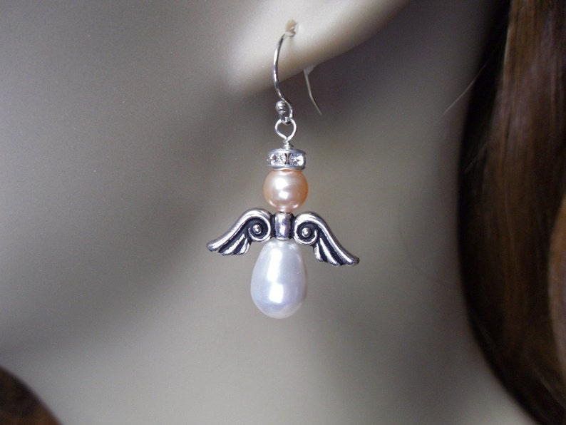 May include: A silver-toned angel earring with a peach-colored bead head, a white pear-shaped pearl body, and silver wings. The earring is suspended from a silver hook, showcasing a delicate and elegant design.