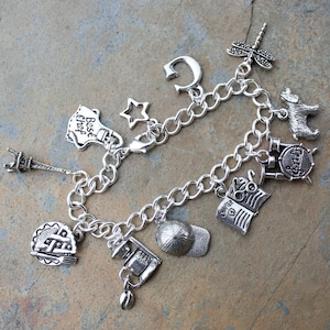 May include: Silver charm bracelet featuring a variety of charms, including a star, a letter C, a chef's hat with the text "Best Chef", a book, a dragonfly, an Eiffel Tower, a coffee pot, a baseball cap, and a musical instrument.