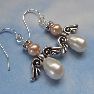 May include: A pair of angel-themed earrings. Each earring features a silver-tone metal angel with pearl-shaped white beads and peach-colored beads. The earrings have silver-tone ear wires and are set against a light blue background.