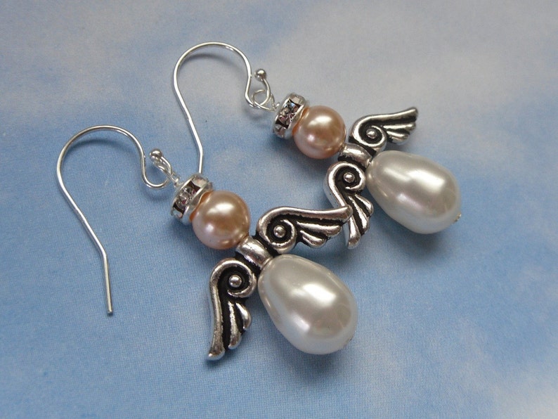 May include: A pair of angel-themed earrings. Each earring features a silver-tone metal angel with pearl-shaped white beads and peach-colored beads. The earrings have silver-tone ear wires and are set against a light blue background.