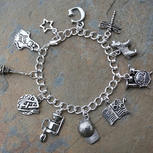 May include: A silver charm bracelet featuring a variety of charms, including a star, a chef's hat, a book, a coffee pot, a baseball cap, an Eiffel Tower, a dragonfly, a drum set, and a dog.