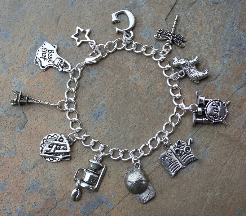 May include: A silver charm bracelet featuring a variety of charms, including a star, a chef's hat, a book, a coffee pot, a baseball cap, an Eiffel Tower, a dragonfly, a drum set, and a dog.