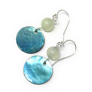 May include: Dangle earrings with iridescent blue-green circular pendants and pale green spherical beads. The earrings are suspended from silver-coloured ear wires. The design is delicate and elegant. The earrings are set against a white background.