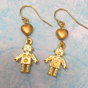 May include: Gold robot earrings with a gold heart charm. The robot is stylized with a round head and a body with three circles.