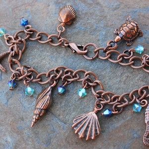 Peut inclure: Un bracelet à breloques cuivré avec des motifs marins. Il comprend une étoile de mer, un hippocampe, des coquillages et une tortue. Orné de perles de cristal bleues et turquoise, c'est un accessoire sur le thème nautique.