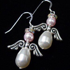 May include: A pair of silver-toned angel earrings with pink and white pearlescent beads. Each earring features a small pink bead at the top, a larger white bead, and silver wings. The earrings have a hook closure.